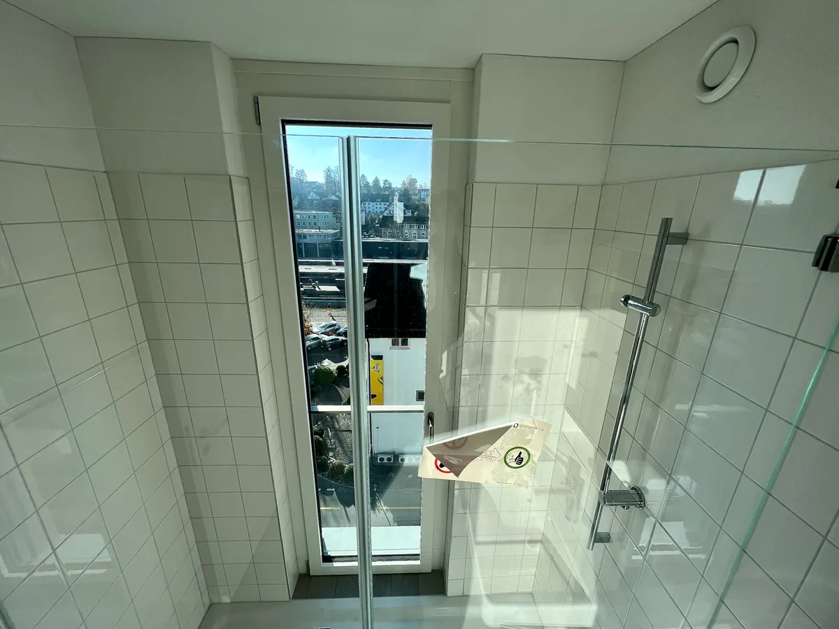 Man sieht eine Dusche, in die ein bodenlanges Fenster eingebaut ist, mit Blick auf eine viel befahrene Strasse. 